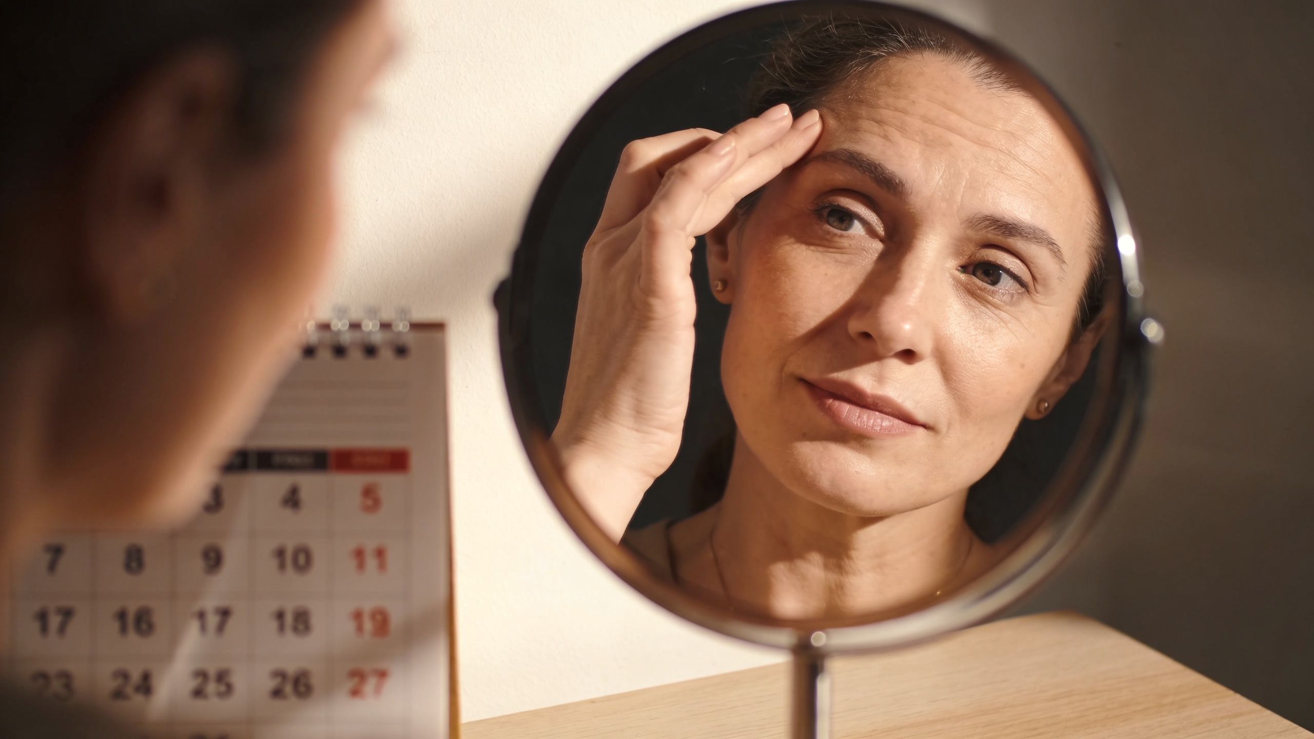 A middle-aged woman looking at her aging skin in a circular mirror next to a calendar.